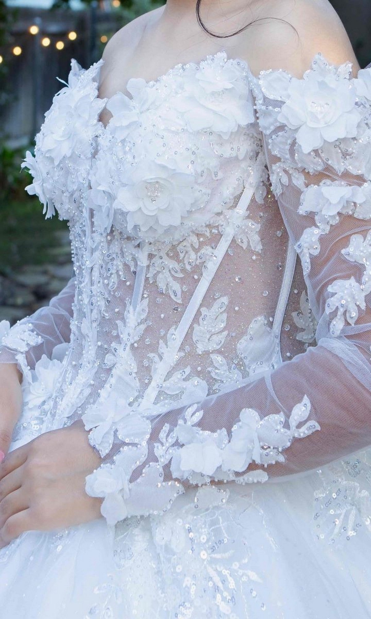 A person wearing the Cinderella Couture 8325J, a long formal white sheer off-shoulder ball gown adorned with floral appliqués, beadwork, and glitter tulle, featuring a delicate corset bodice, photographed outdoors.